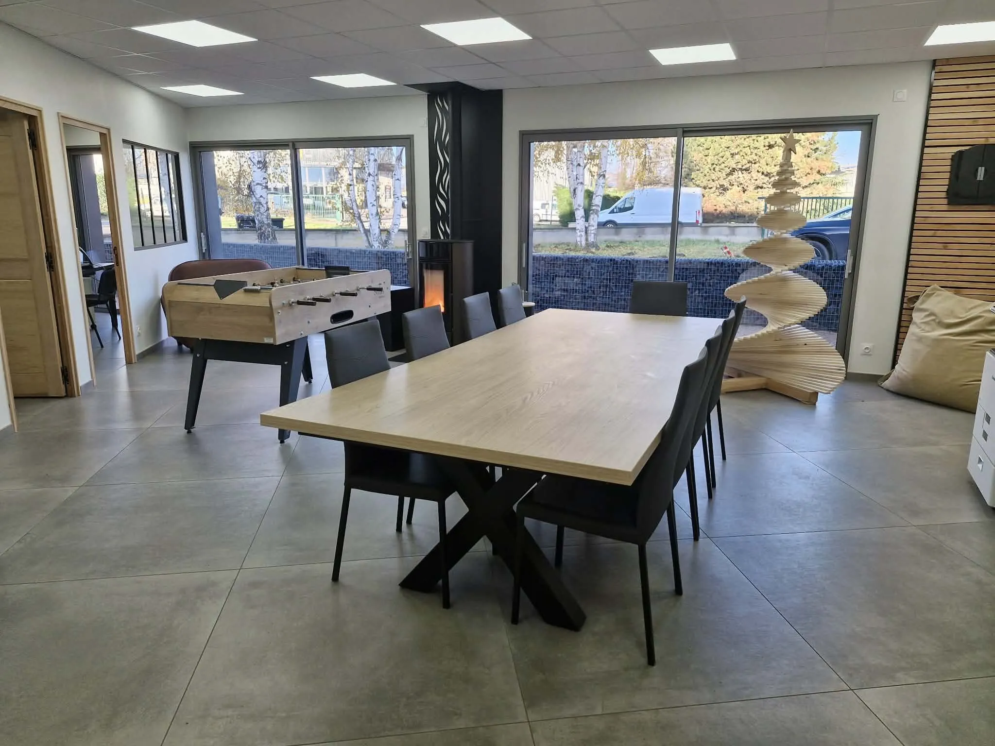 Salle commune lumineuse avec une grande table en bois à piétement en X, chaises noires et baby-foot en arrière-plan