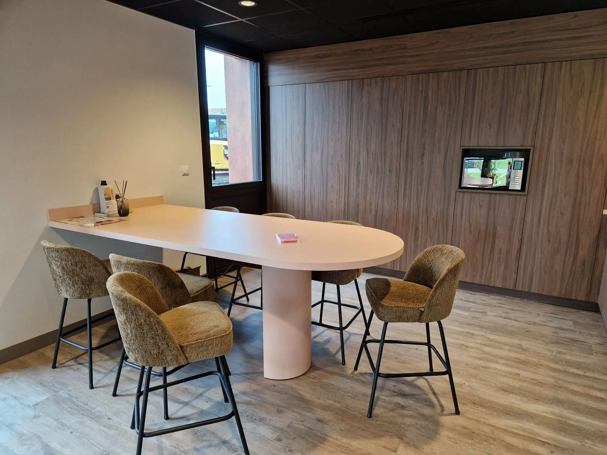 Salle de pause aménagée pour AXA, avec une table haute aux bords arrondis, six chaises hautes et un mur habillé de bois qui intègre un écran